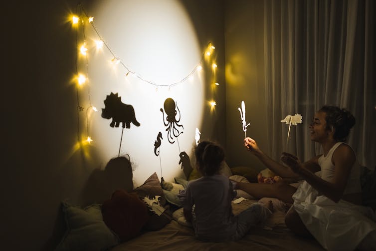 Children and a woman hold stick figures of animals on a lighted circle on the wall in a dark room.