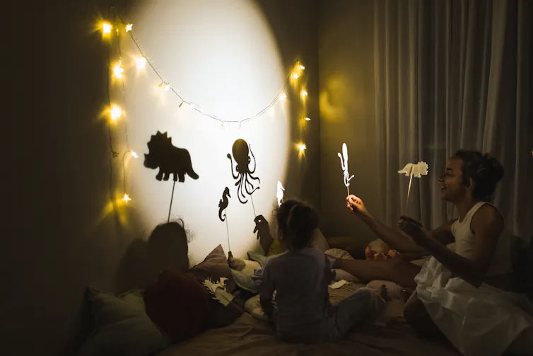 Children and a woman hold stick figures of animals against a lit-up circle on a wall in a dark room.