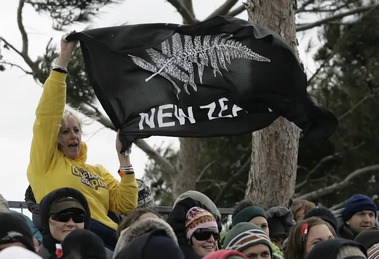 A woman in a yellow hoodie waves a silver fern flag.