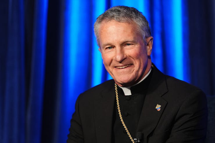 A man with gray hair and a black suit with a clerical collar smiles as he sits in front of a bright blue curtain.