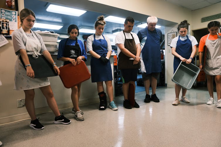 Seven people wearing aprons stand with their heads bowed