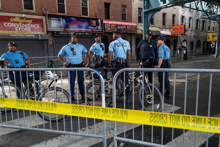 A group of uniformed police stand on an empty commercial street behind metal gate and yellow police tape
