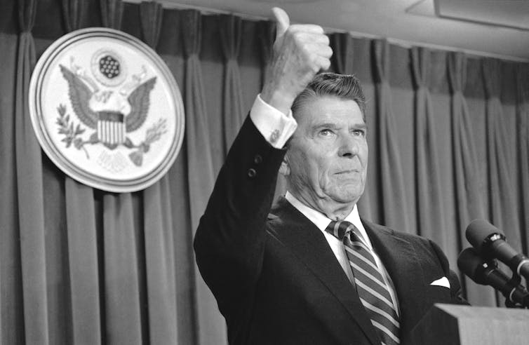 A man in a striped suit and tie stands at the podium in front of the presidential seal, raising his arm in an upward gesture.