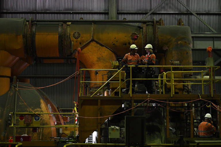Two men wearing yellow hard hats stand in a dim machinery room in a power station