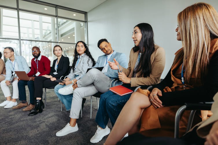 People sit on chairs in a partial circle, and a woman speaks while others look at her