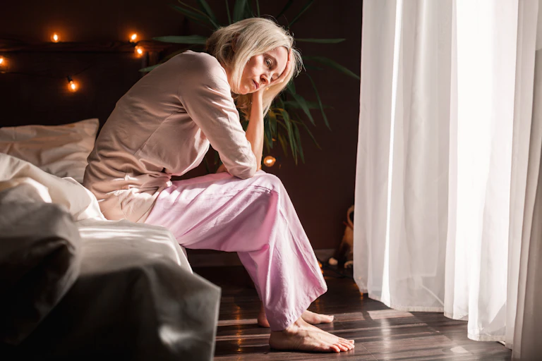 A tired woman sits on the edge of her bed, resting her head in her hand.