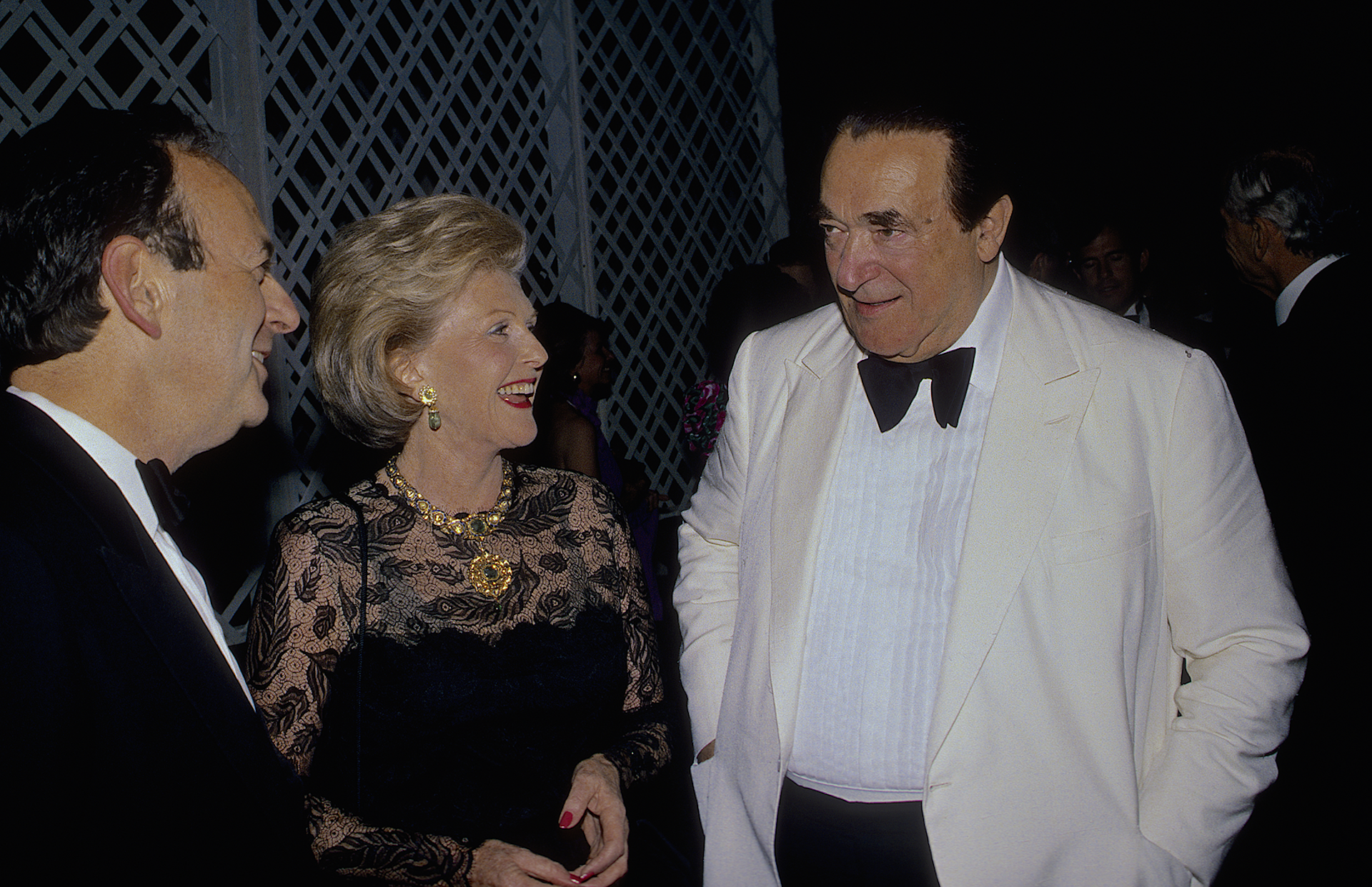 A large man in a white dinner jacket talks with a middle aged woman in evening dress and another man in a black dinner jacket.
