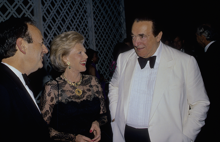 A large man in a white dinner jacket talks with a middle aged woman in evening dress and another man in a black dinner jacket.