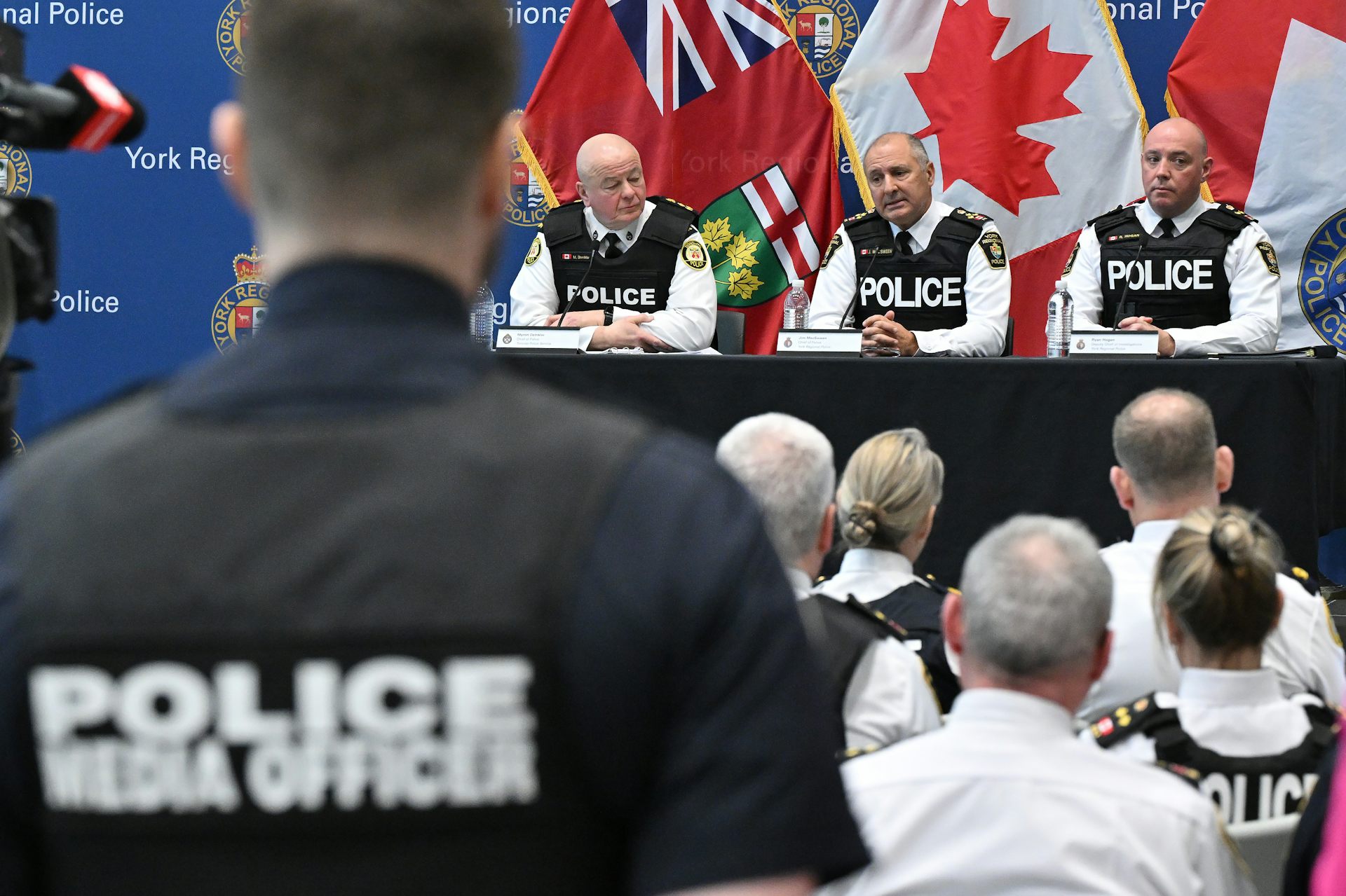 Three men wearing bullet-proof vests at a news conference with a man in the foreground photographed from behind wearing a shirt that reads Police Media Officer.