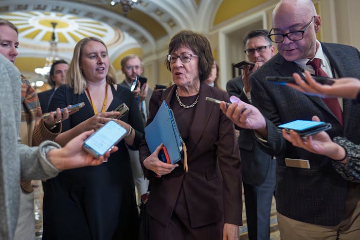 Sen. Susan Collins, holding a portfolio, speaks as she's surrounded by reporters on Capitol Hill.