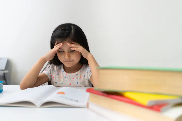 A young girl with dark hair sits with her hands on her head and looks at an open book on a desk.