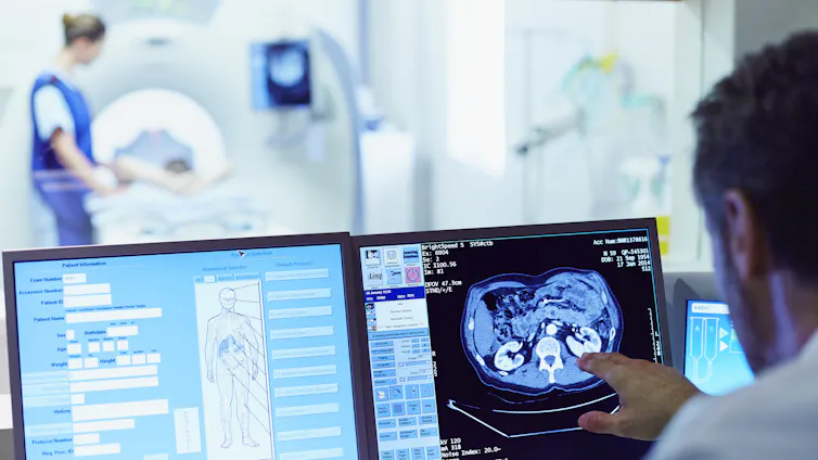 A doctor running a CT scan from a control room, with patient and nurse in the background