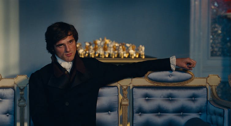 Jacob Elordi as Heathcliff: a darkly brooding, well-dressed man sits on an ornate blue couch with gold, arm outstretched