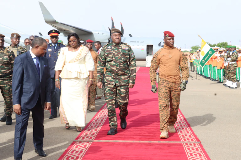 Two men in military uniform walk on a red carpet with an aeroplane in the background