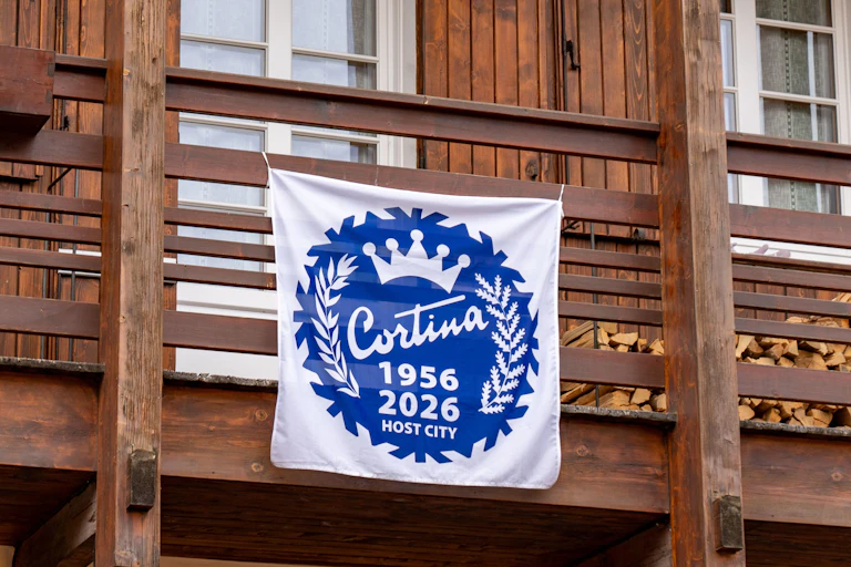 A flag draped over the side of a chalet in Cortina d'Ampezzo, reading Cortina 1956-2026.