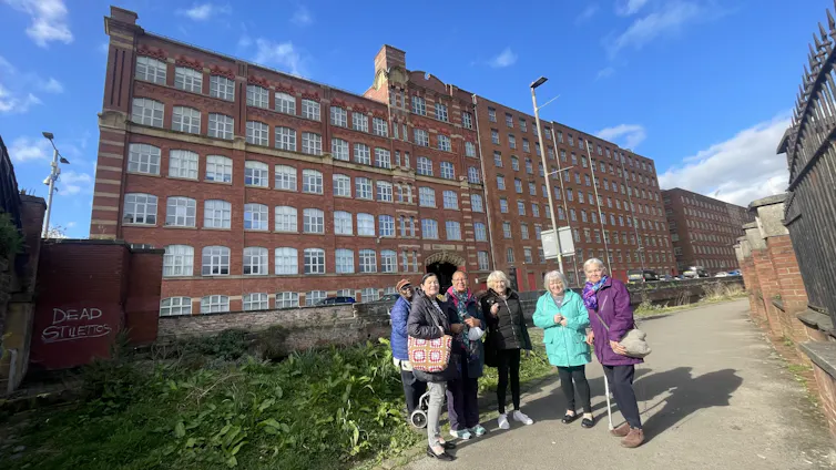 group of older artists stand in front of old red brick mill building