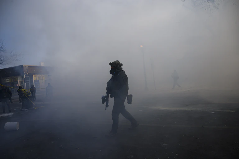 A man dressed in military gear and holding a rifle is surrounded by tear gas.