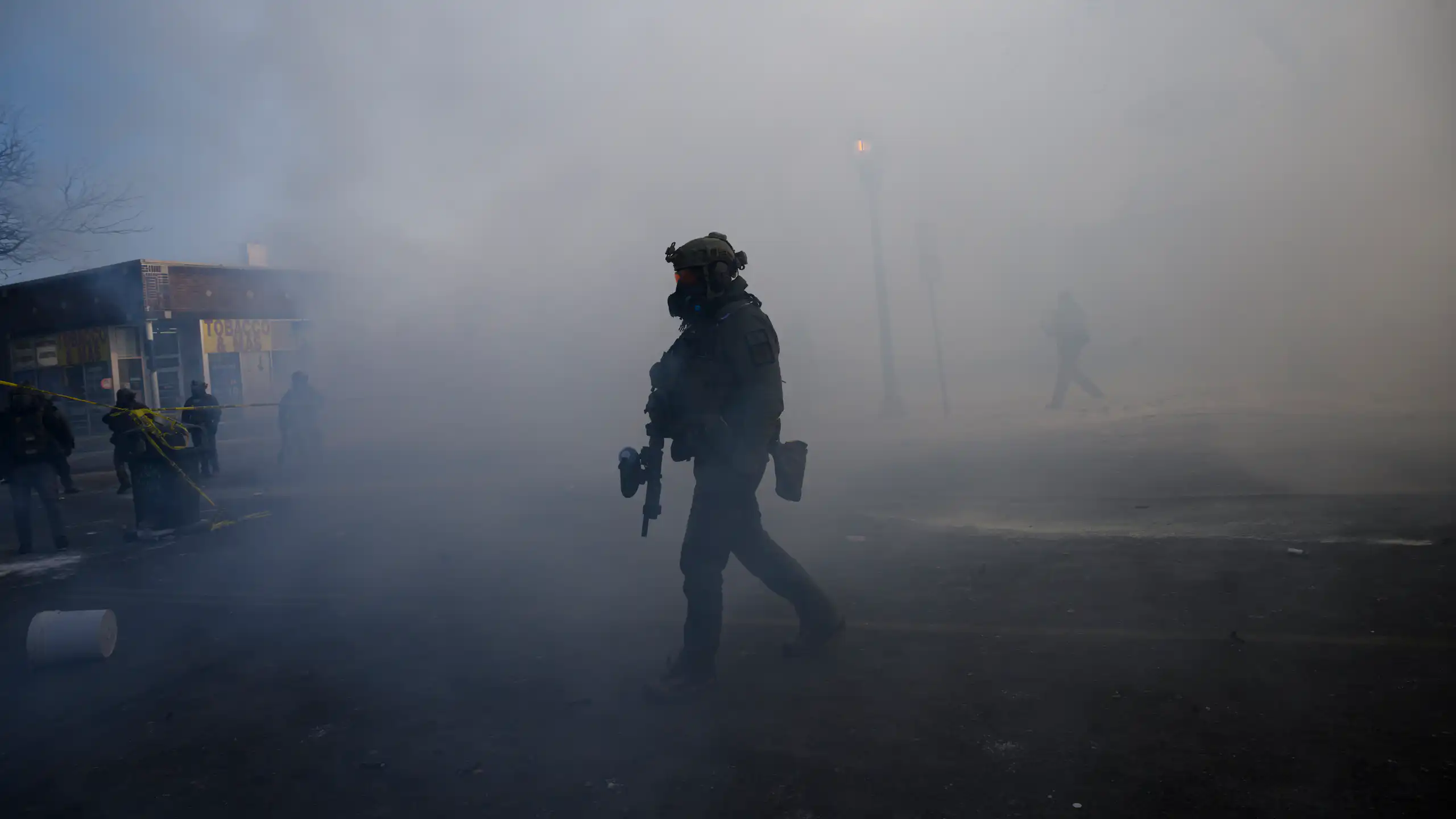 A man dressed in military gear and holding a rifle is surrounded by tear gas.
