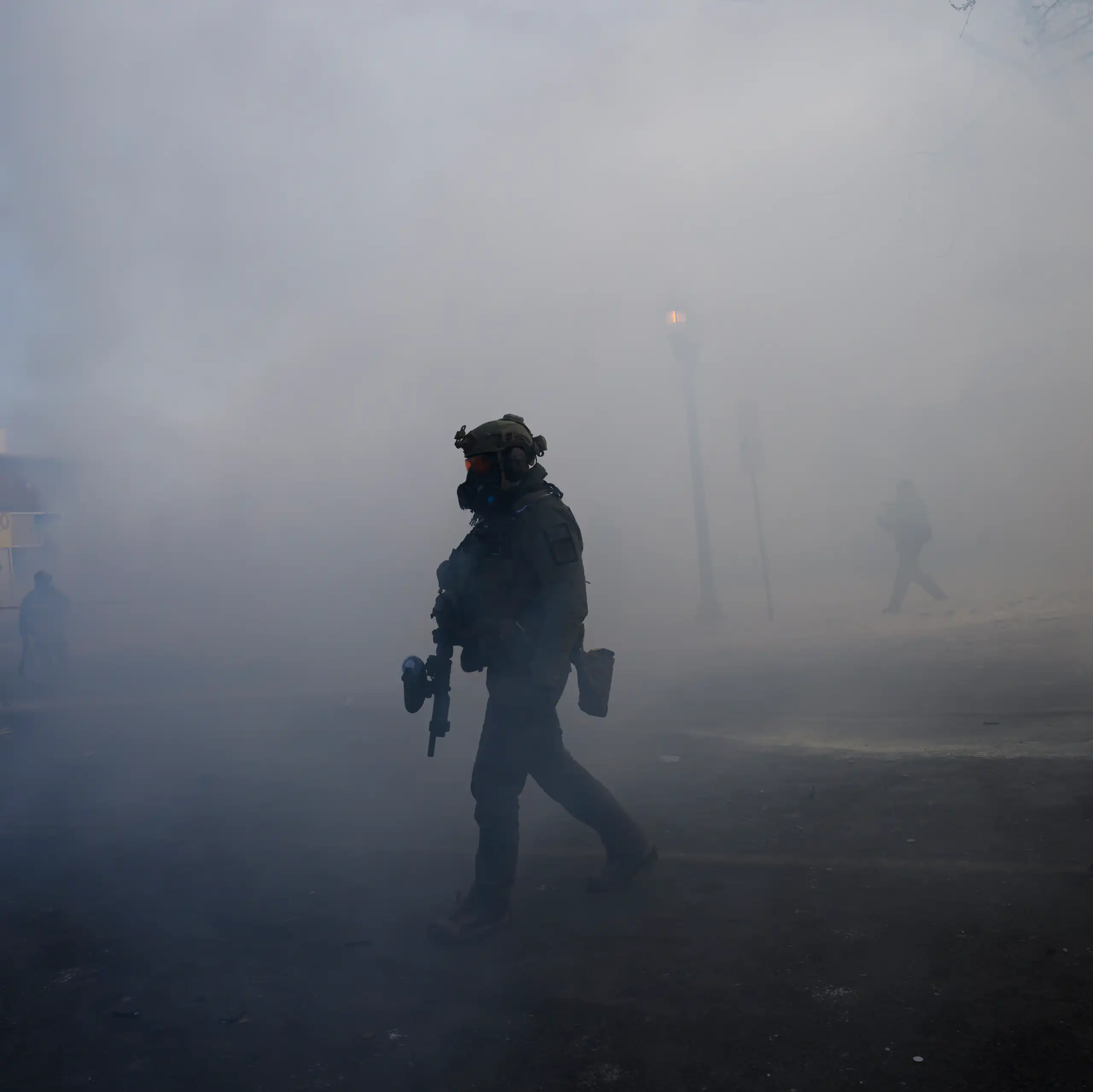 A man dressed in military gear and holding a rifle is surrounded by tear gas.