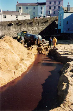 people on beach with stream of black oil, tanker in distance