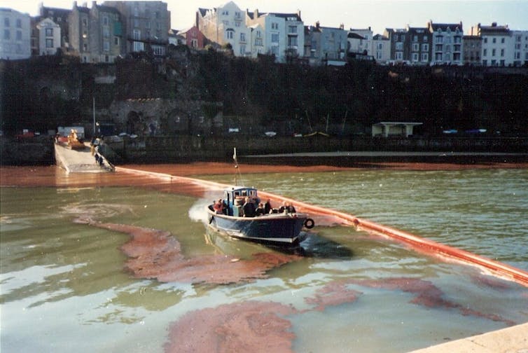 river with dark oil, brown boom stretches across width with boat, houses in background