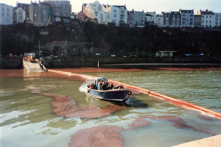 river with dark oil, brown boom stretches across width with boat, houses in background
