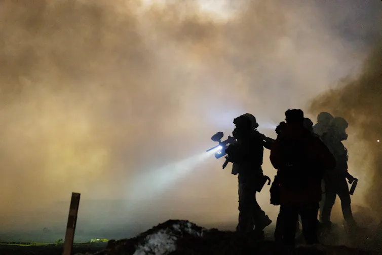 Men dressed in military gear and holding rifles are surrounded by tear gas at night.