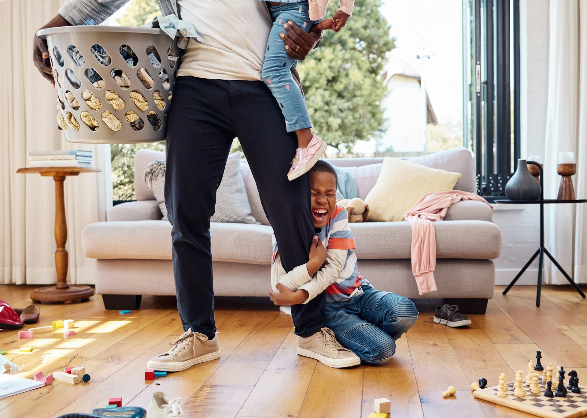 A boy in a home with a parent holding a laundry bin and a younger sibling is holding the parent's leg with an excited look on his face. 