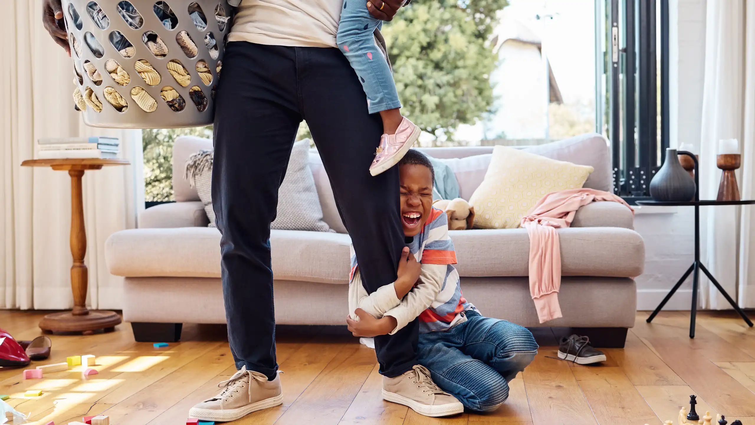 A boy in a home with a parent holding a laundry bin and a younger sibling is holding the parent's leg with an excited look on his face.