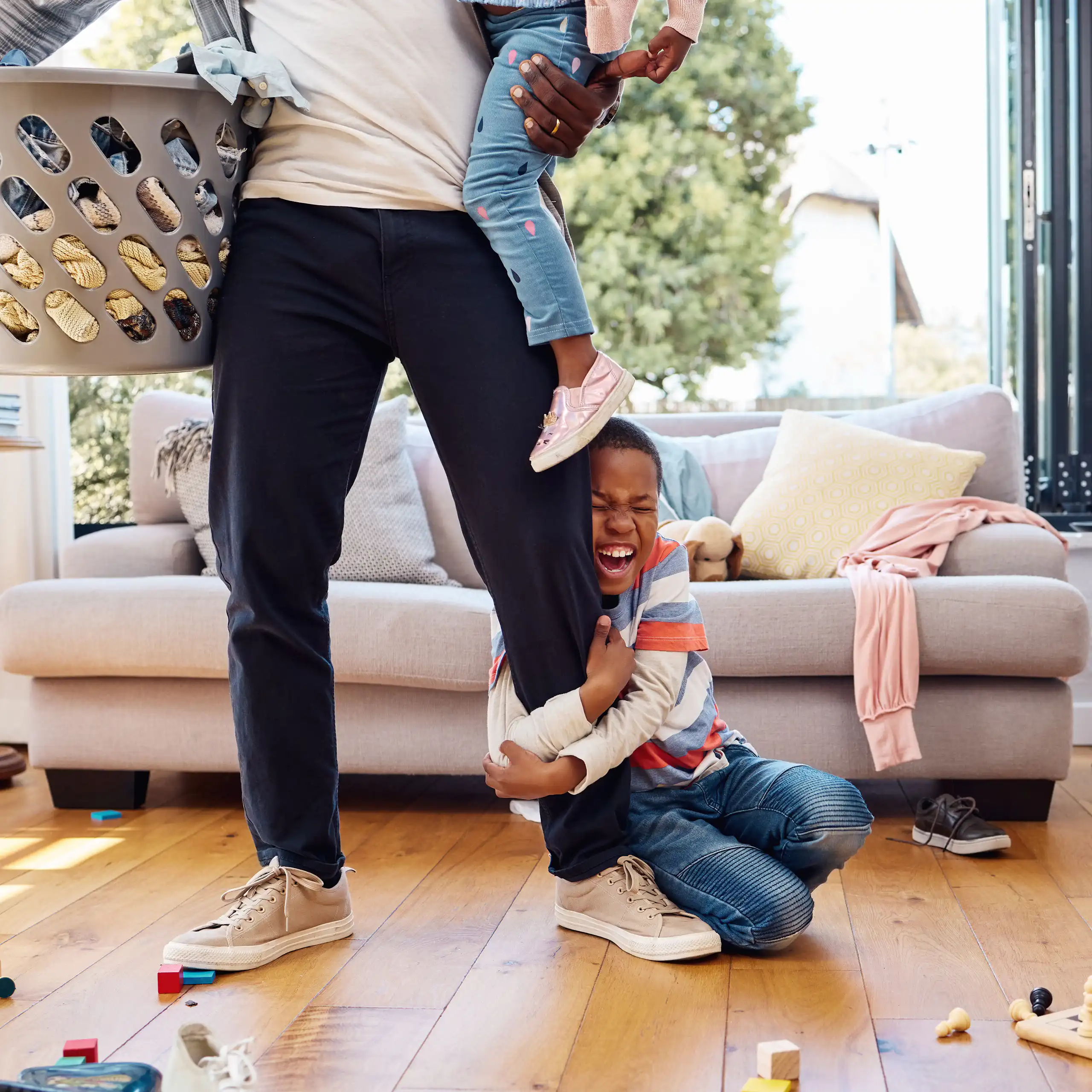 A boy in a home with a parent holding a laundry bin and a younger sibling is holding the parent's leg with an excited look on his face.