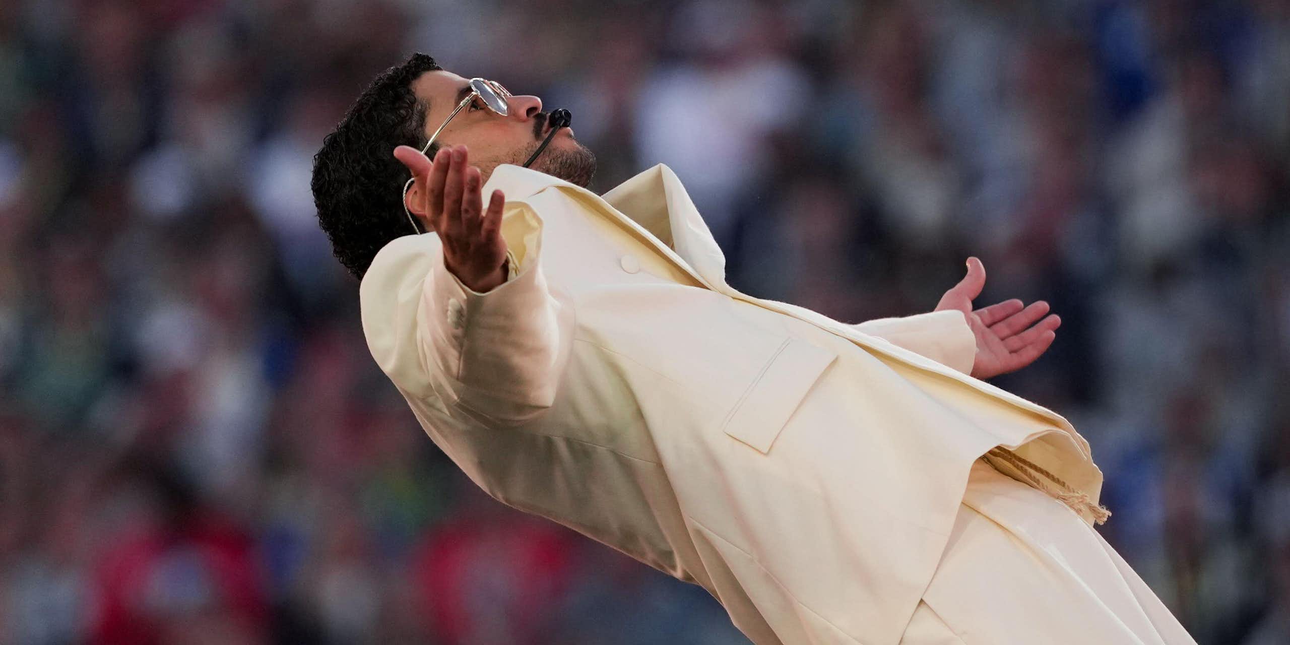 Um homem vestido com um terno creme e óculos escuros absorve a adoração da multidão em um show no palco.