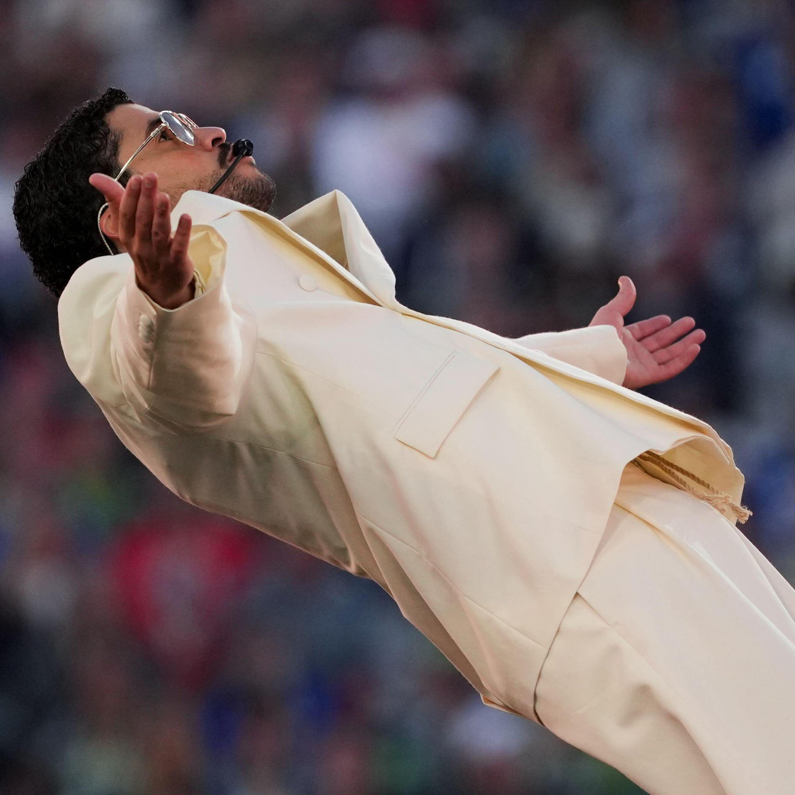 Um homem vestido com um terno creme e óculos escuros absorve a adoração da multidão em um show no palco.