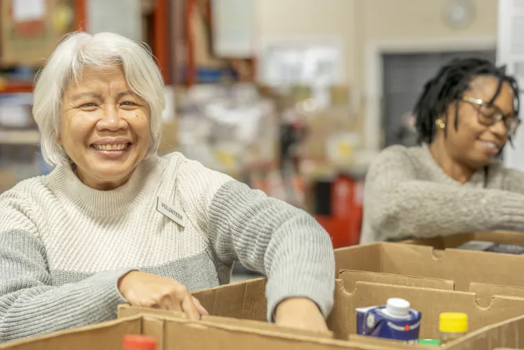 An Asian woman with white hair smiles as she puts cans of food into boxes at a food pantry, standing beside a younger Black woman.