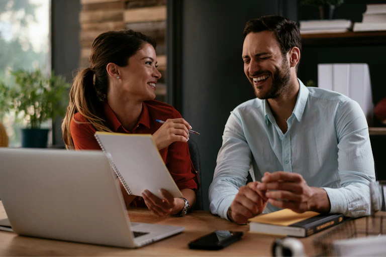 Two people talking and smiling while sitting at a desk doing work