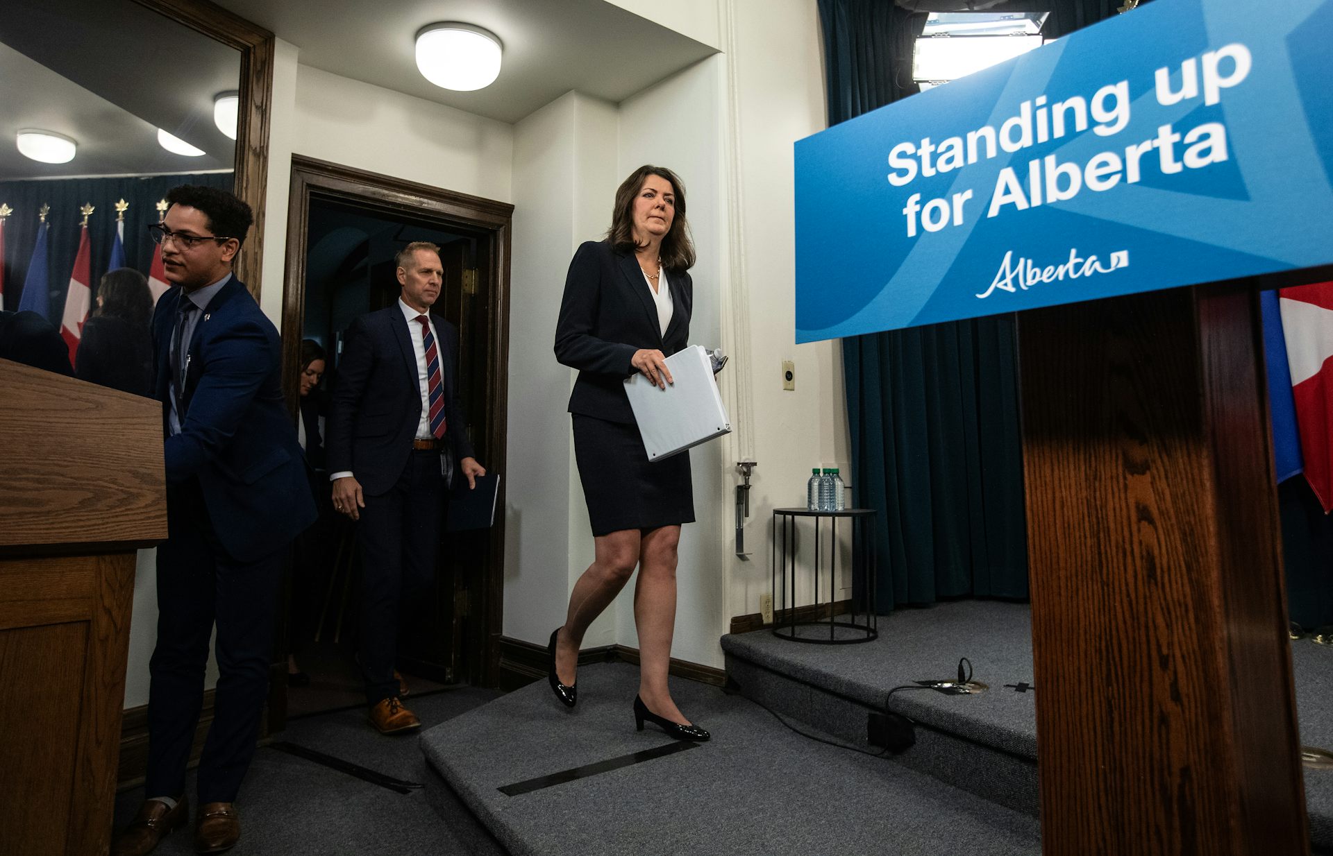 A dark-haired woman walks towards a podium and a sign that reads Standing up for Alberta.