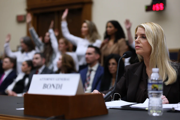 US attorney-general, Pam Bondi, sits at a desk behind a name plate. IN the background is a group of women with their hands raised.