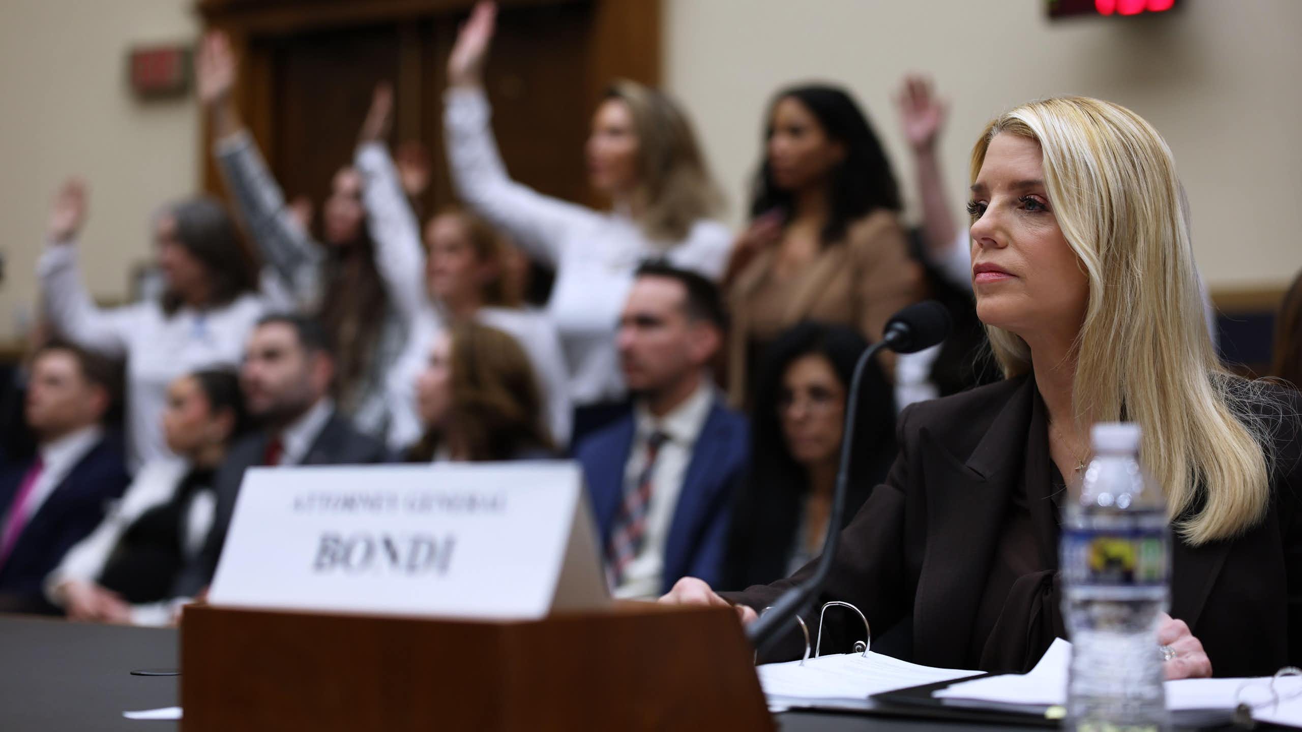 US attorney-general, Pam Bondi, sits at a desk behind a name plate. IN the background is a group of women with their hands raised.