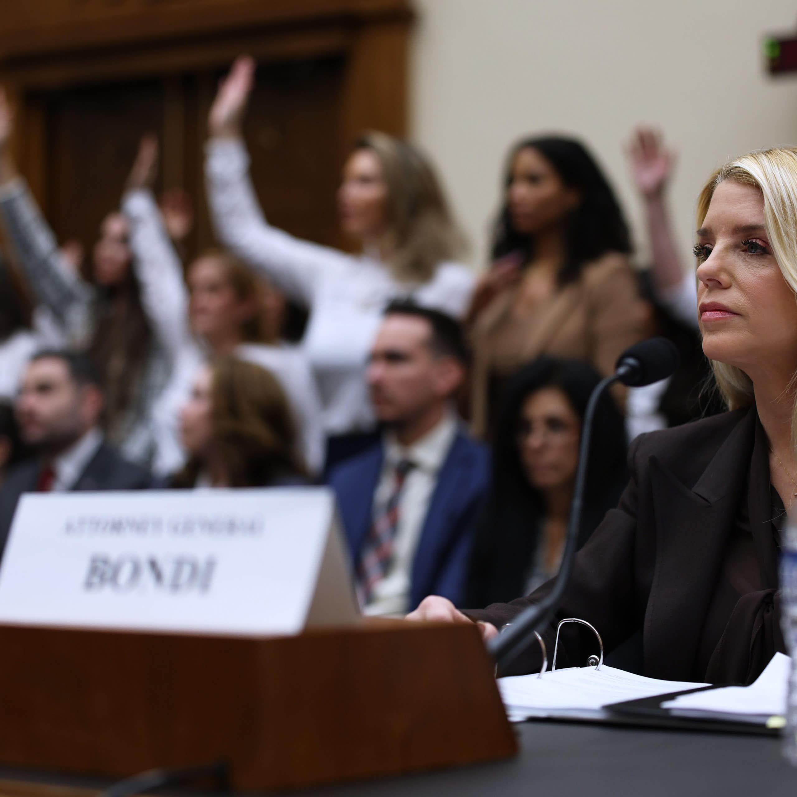 US attorney-general, Pam Bondi, sits at a desk behind a name plate. IN the background is a group of women with their hands raised.
