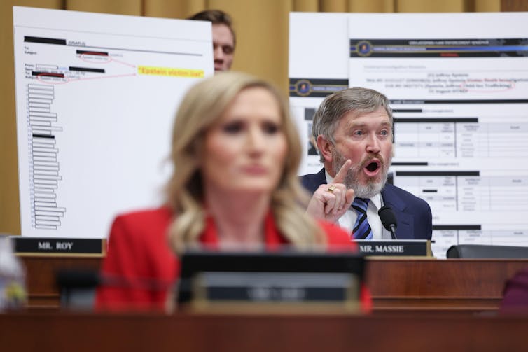 A man speaks and points. Behind him are projections of documents with some details blacked out. In the foreground a woman sits at her compujter.