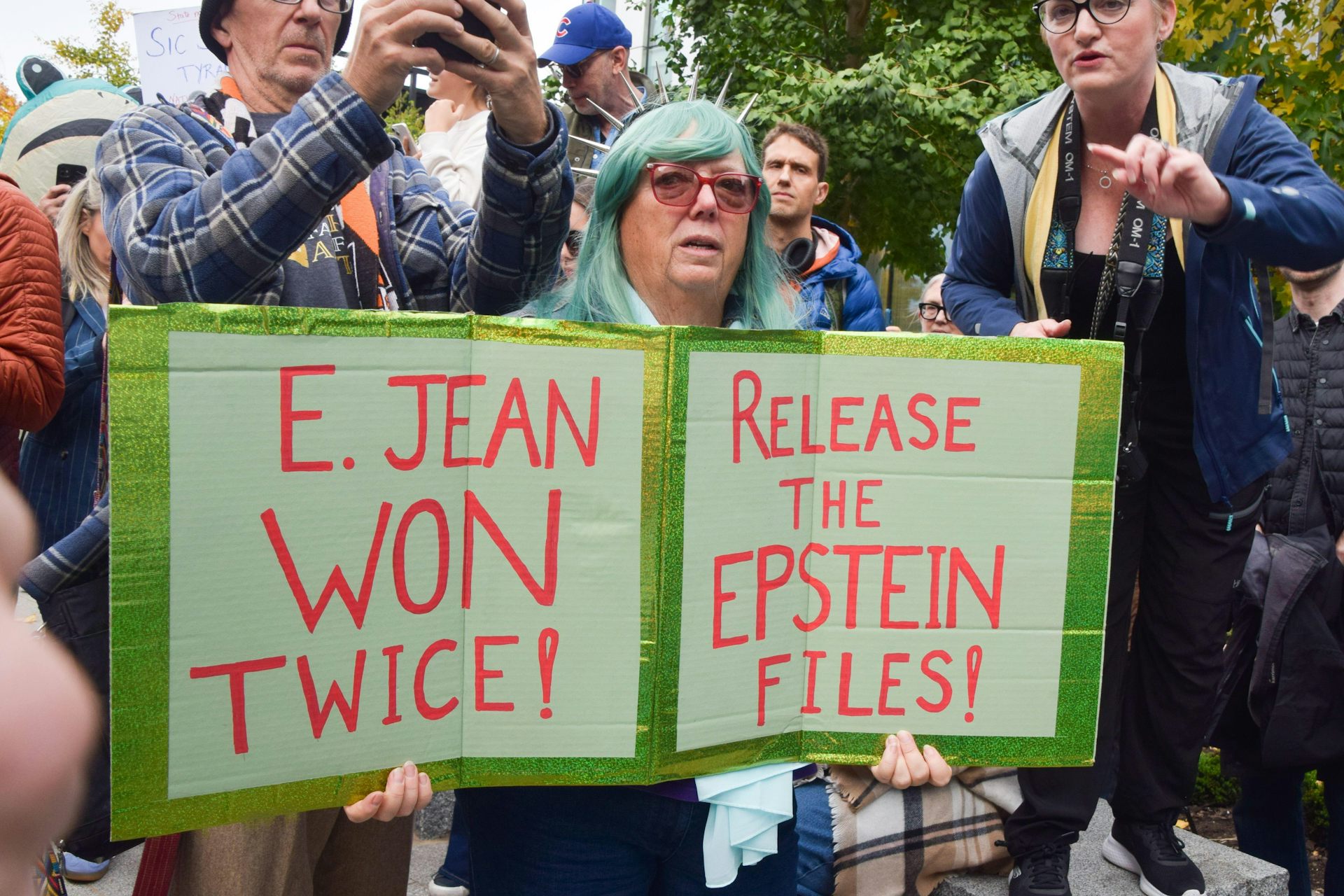 A woman stands at a protest rally with two signs. One says: E Jean won twice. The othe rsays: Release the Epstein files.