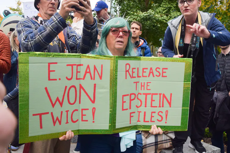 A woman stands at a protest rally with two signs. One says: E Jean won twice. The othe rsays: Release the Epstein files.