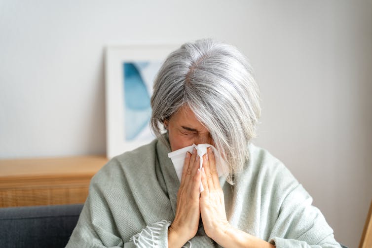 An elderly woman sneezes into a tissue