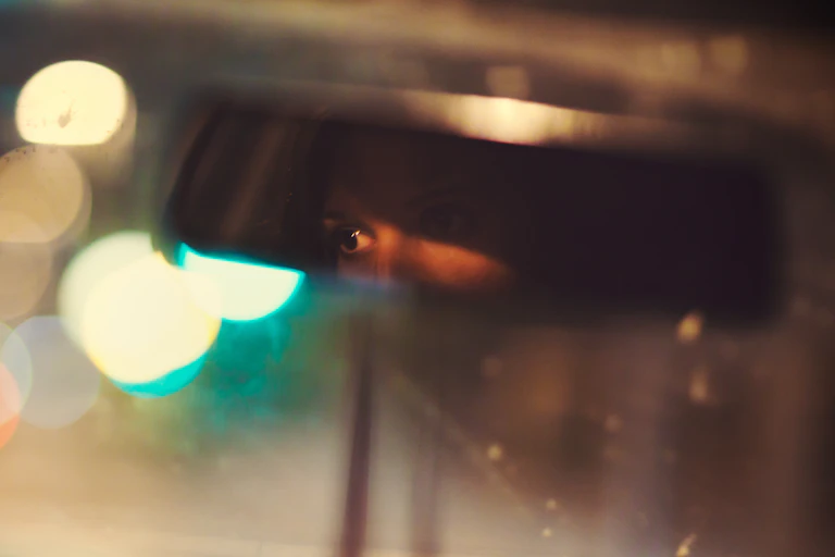 A woman drives a car at night with her eyes visible through the rearview mirror.