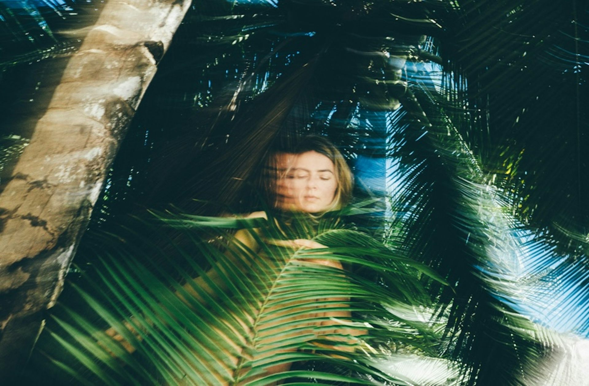A women with closed eyes is partially visible amidst the dense canopy of palm fronds.