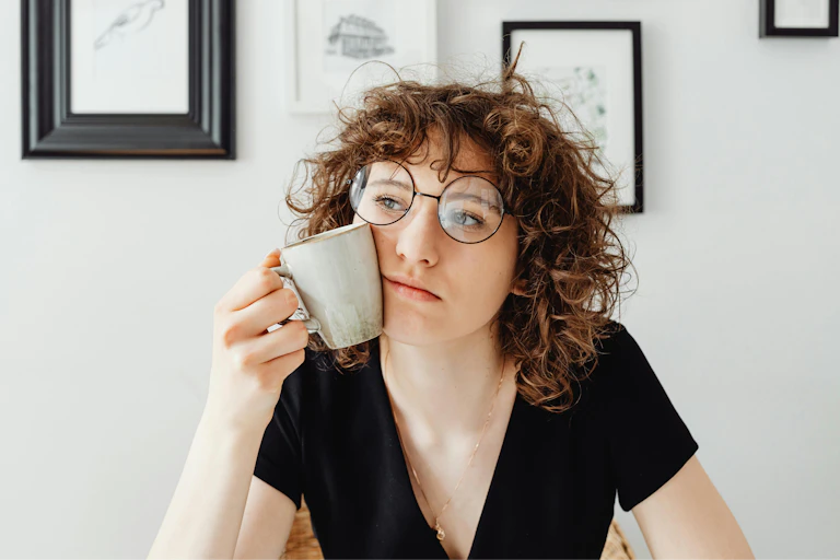 Mulher com cabelo encaracolado e óculos segura uma xícara de café contra o rosto enquanto pensa.