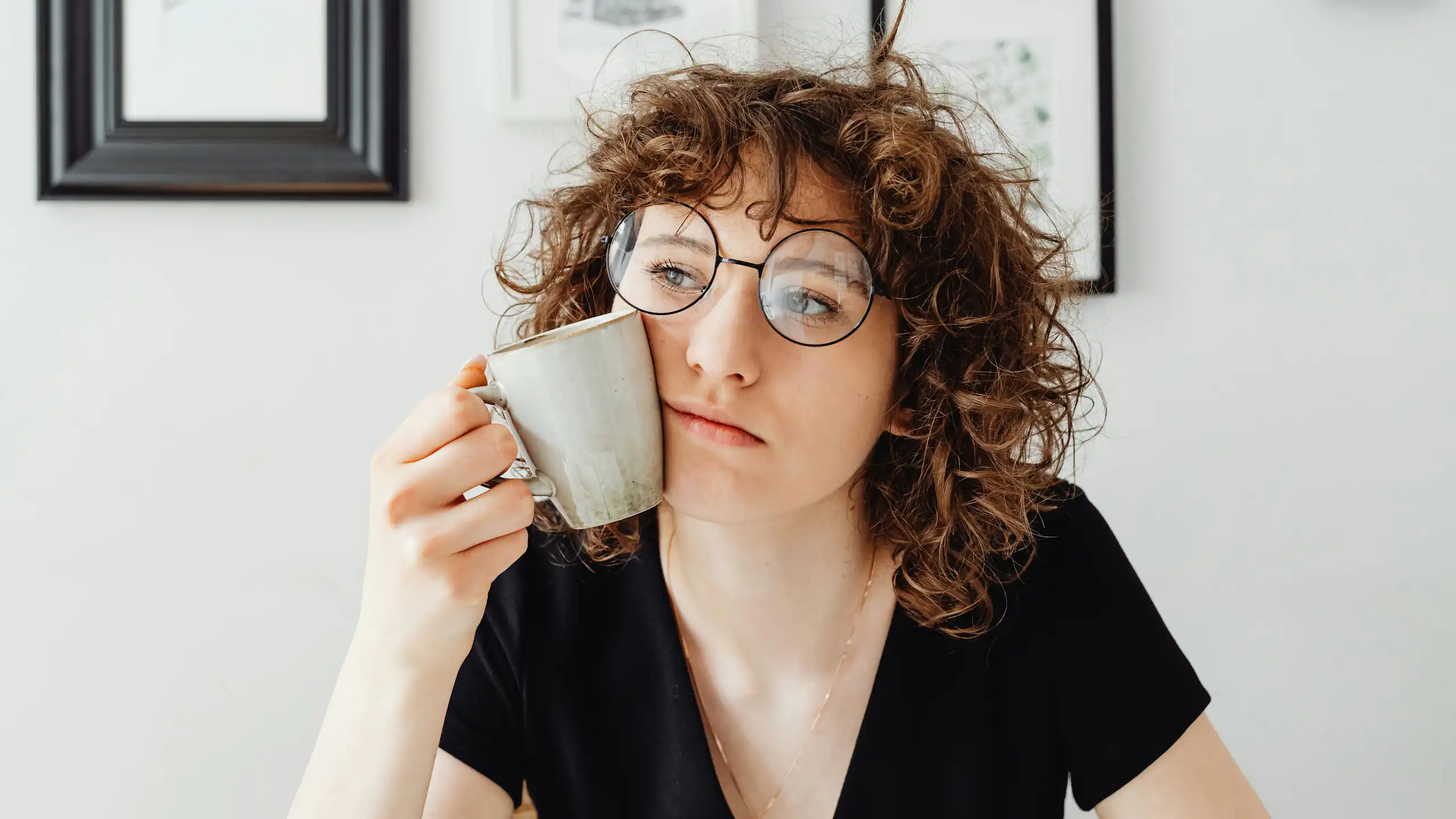 Mulher com cabelo encaracolado e óculos segura uma xícara de café contra o rosto enquanto pensa.
