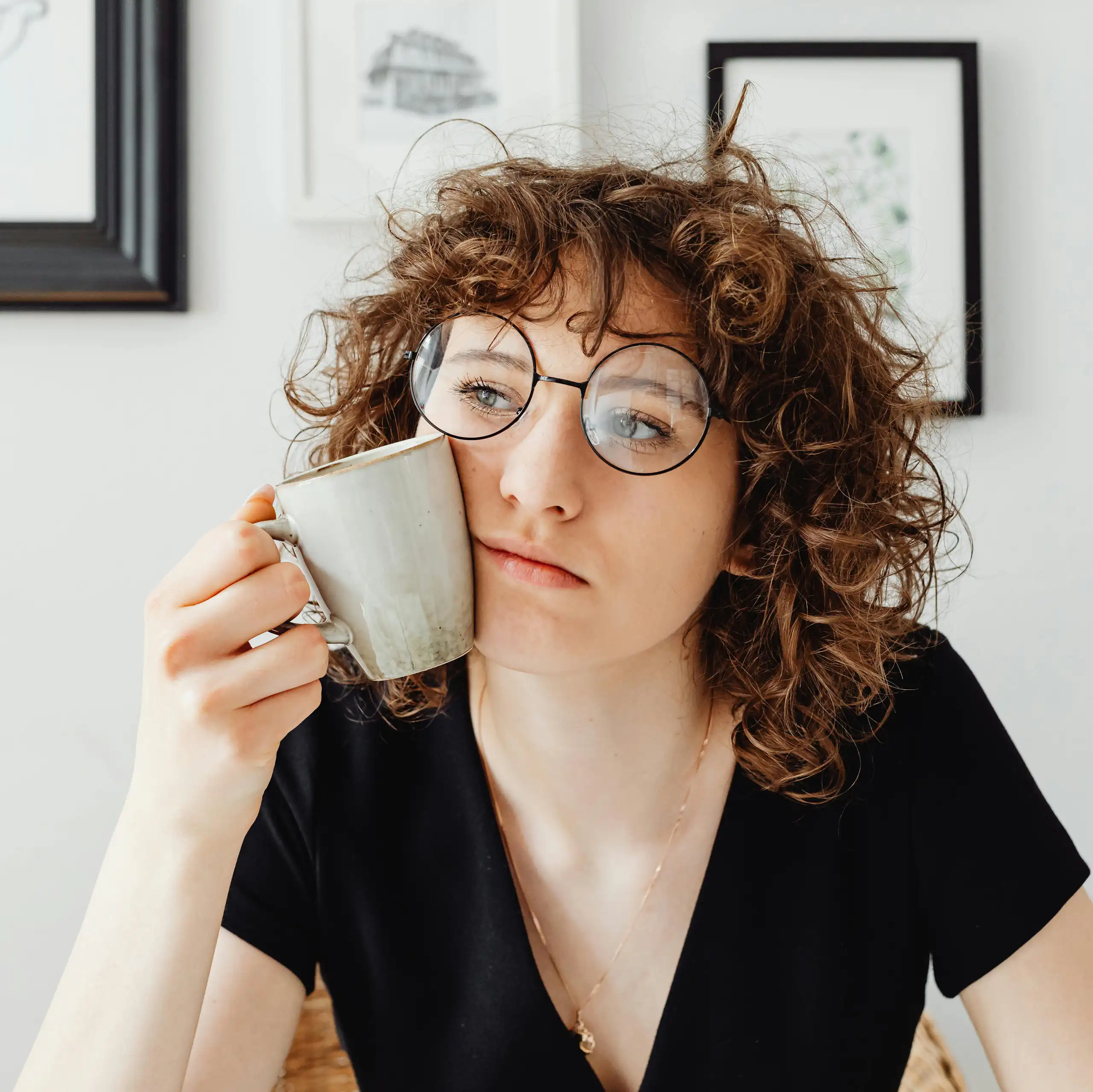Mulher com cabelo encaracolado e óculos segura uma xícara de café contra o rosto enquanto pensa.