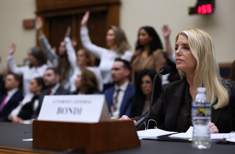 US attorney-general, Pam Bondi, sits at a desk behind a name plate. IN the background is a group of women with their hands raised.