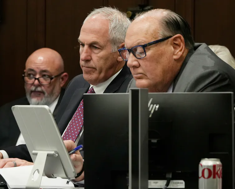 Two men in suits sit at a table, with another man in the background.