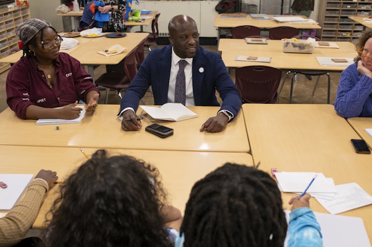 A group sits around tan school tables, looking and talking with each other.